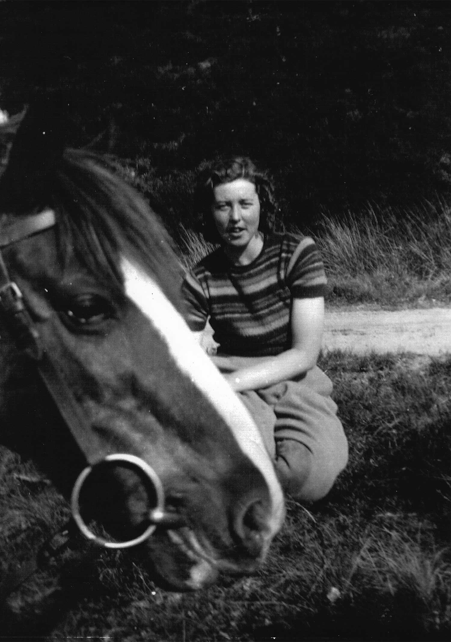A black and white photo of a young woman in a short-sleeved striped shirt and pants, crouching on a lawn. A horse with a white blaze, wearing a bridle, pokes its head into the photo from the left.
