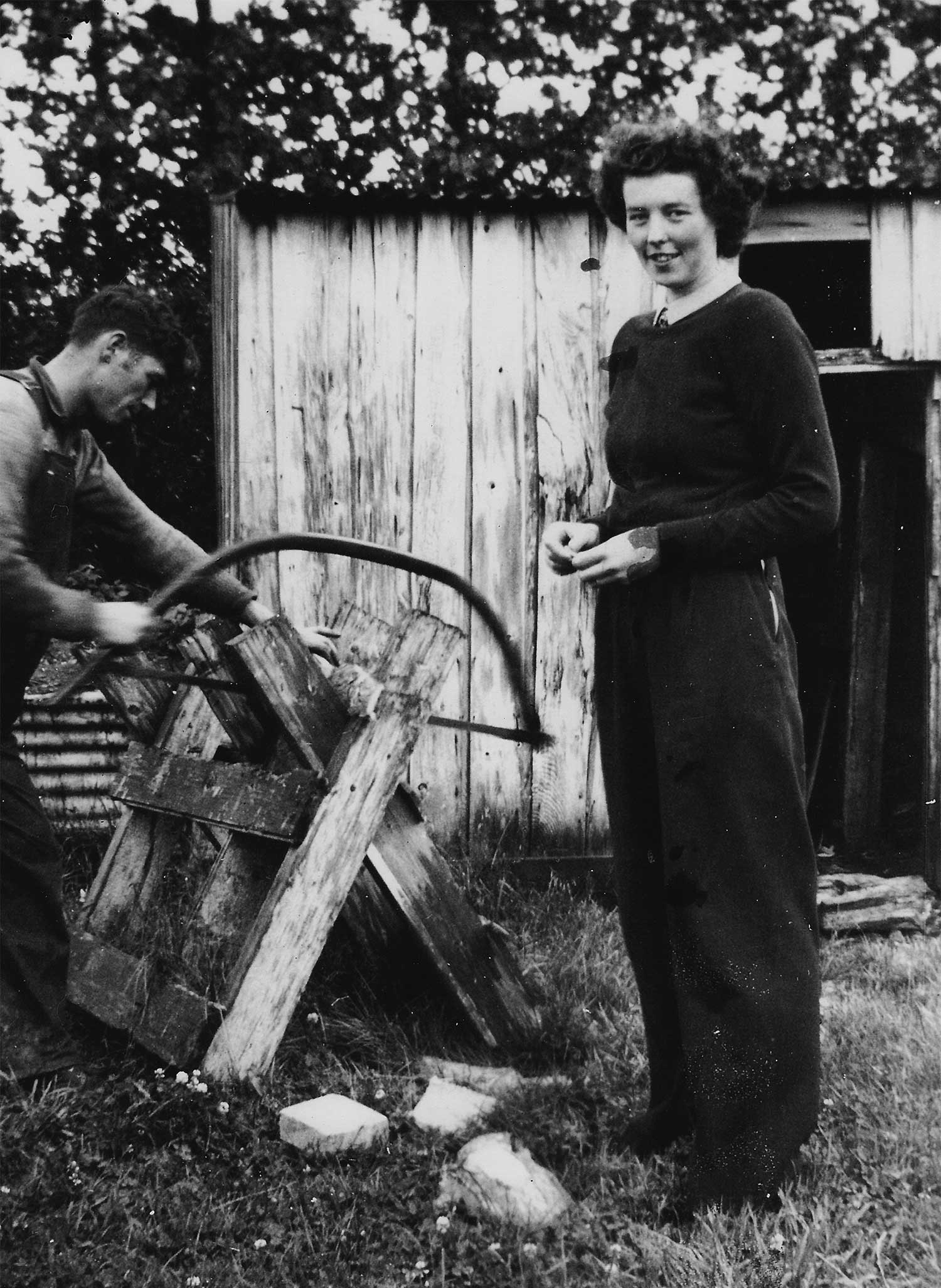 A black and white photograph of a man and a woman outside a shed. He is concentrating on sawing a log on a sawhorse. She wears a dark sweater and pants and smiles at the camera.