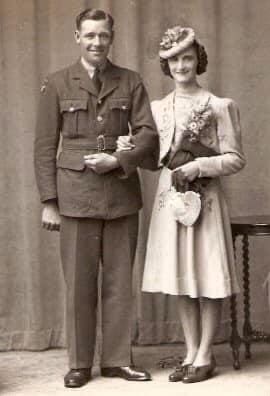 A sepia photograph of a man in uniform and a woman in a light-coloured dress and a round hat sitting tilted and high on her head. They stand arm in arm and smile sweetly at the camera.