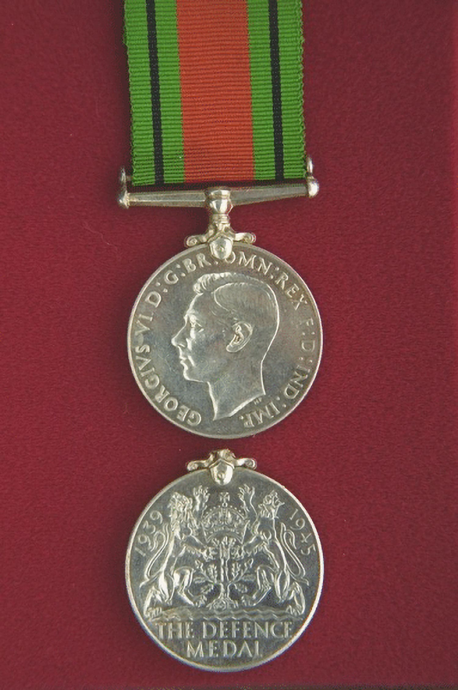 A photograph of a round silver medal suspended from a green and red striped ribbon. The medal shows a profile of King George VI, facing left. Around its edge is an abbreviated Latin engraving. The obverse side of the medal is also displayed. It features the words THE DEFENCE MEDAL below an oak tree topped by the Royal Crown and flanked by a lion and a lioness. The years 1939 and 1945 also appear.