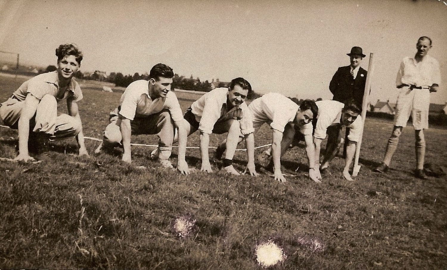 A sepia photograph of five men in shirtsleeves crouched in short grass, taking the starting position for a foot race.