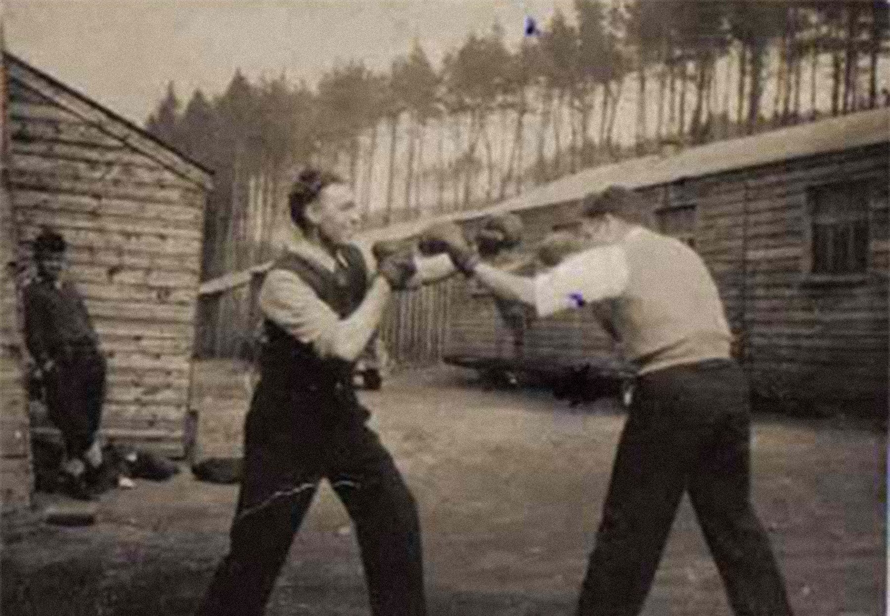 A sepia photograph of two men in shirtsleeves, knitted vests and boxing gloves facing each other and throwing punches. A third man leans against a rough wooden building, watching.