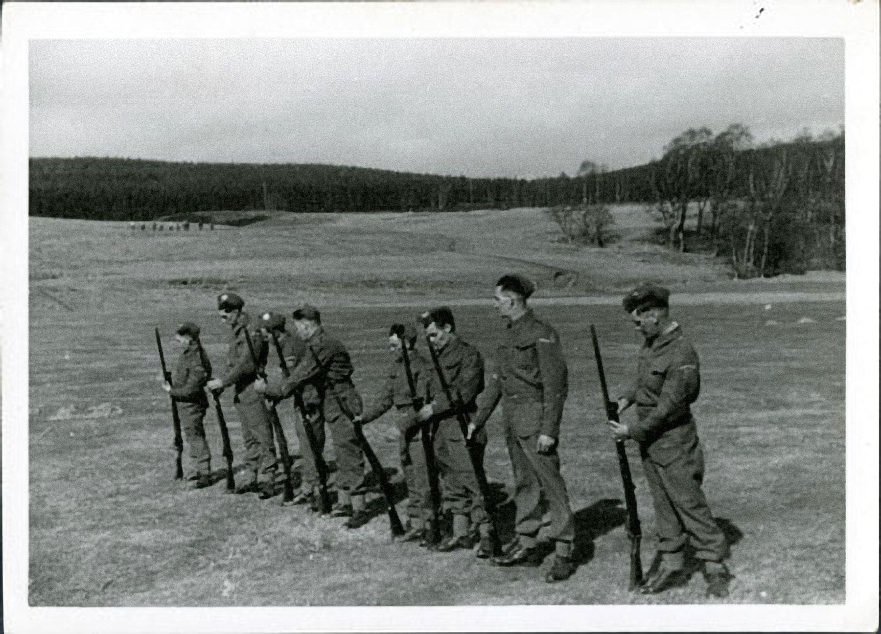 A black and white photograph of eight men in uniform lined up in an open field. They hold their rifles in front of them, butts resting on the ground.