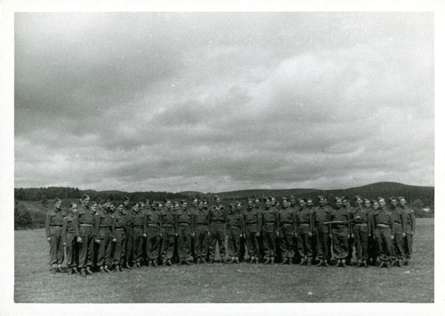A black and white photograph of dozens of men in uniform in an open field under a cloudy sky. They stand at attention in three rows, facing the camera.
