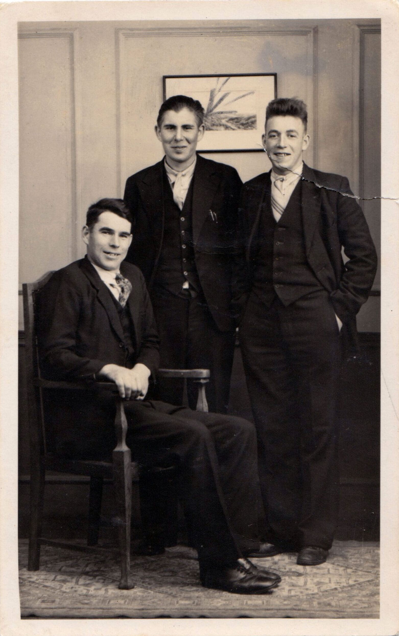 A sepia photograph of three young men in dark suits and ties in a living room or studio. The one on the left is seated, the one on the right stands with his hands in pants pockets. All are smiling at the camera.