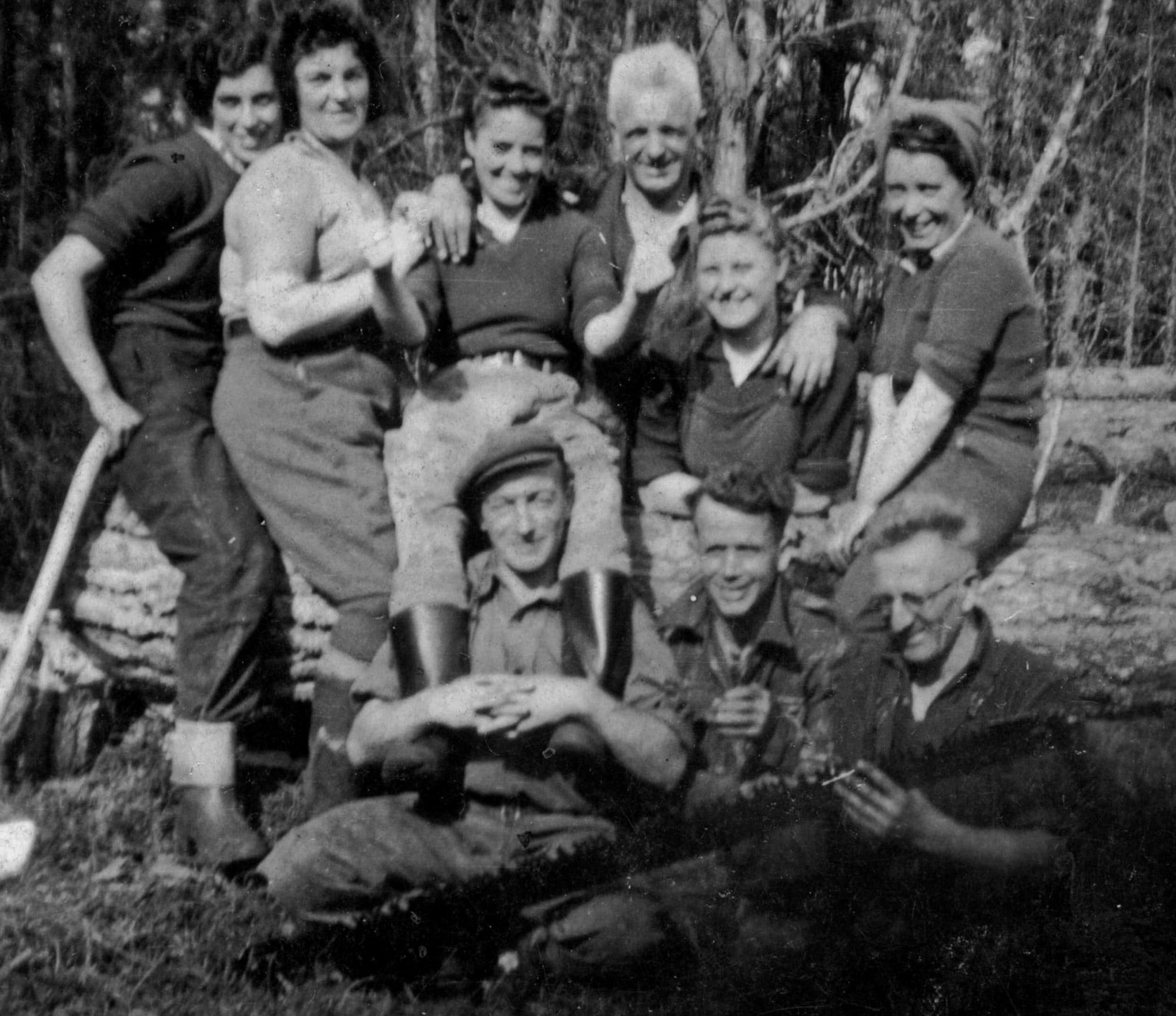 A black and white photograph of a group of women and men in casual clothes sitting on or in front of a felled tree trunk, smiling at the camera.