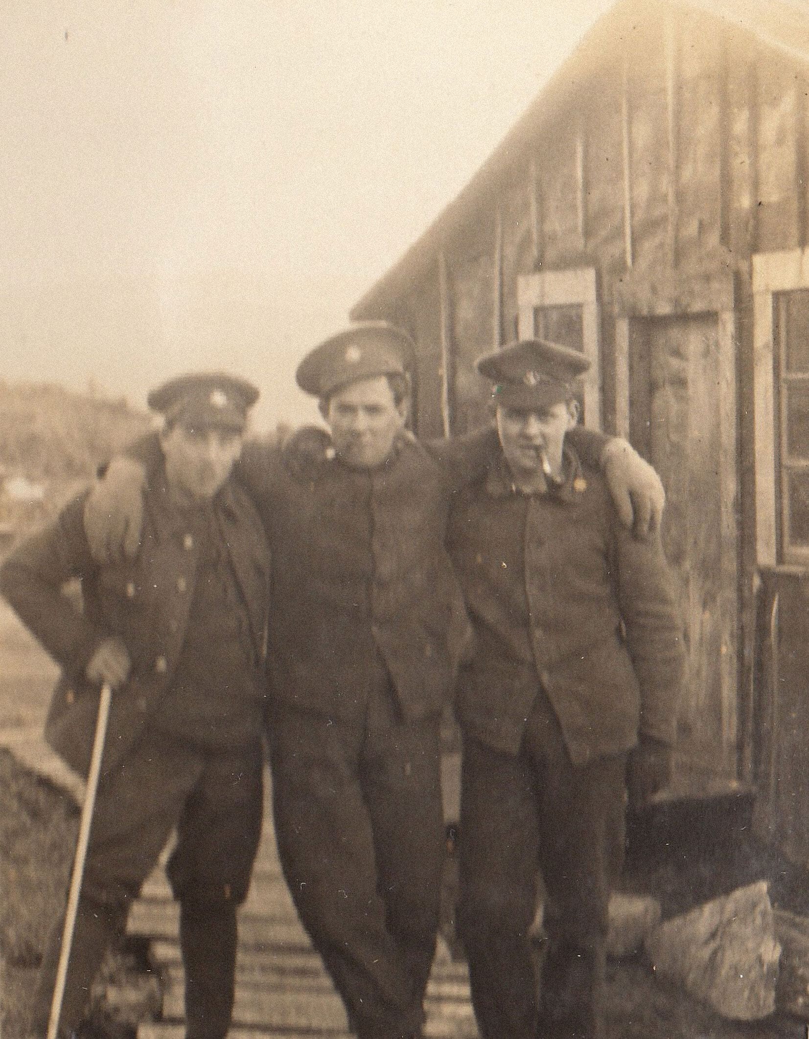 A sepia photograph of three soldiers in uniform standing casually. The man in the centre wears his cap at an angle and drapes his arms over the shoulders of his pals. The man on the left leans on a swagger stick and the man on the right holds a pipe in his mouth.