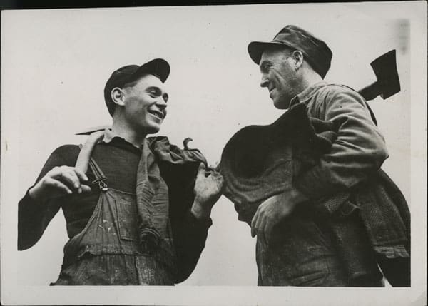 A black and white photograph of two smiling loggers in overalls. They face one another, axes resting over their shoulders. The younger man, jacket hooked on this thumb, is at left. The older man holds his coat under his arm.