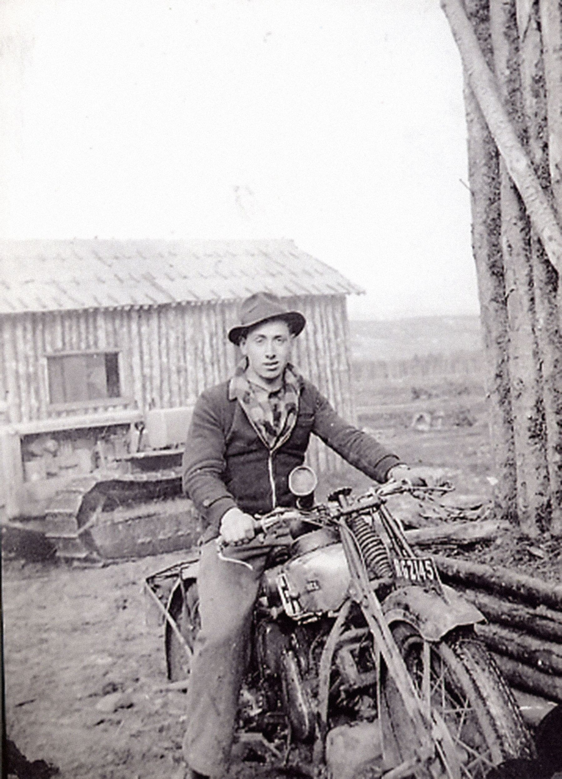 A black and white photograph of a young man in casual clothes sitting astride a motorcycle. Behind him are a Caterpillar tractor (on continuous tracks, not wheels) and a rough log building.