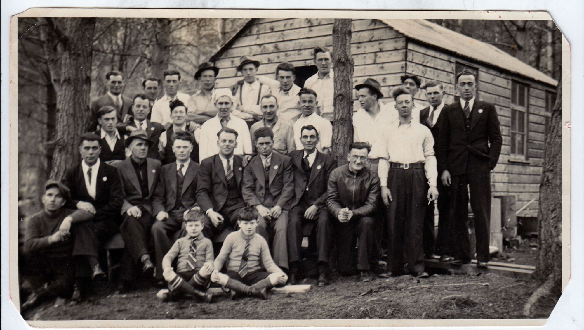 A black and white photograph of a large group of foresters sitting and standing in rows outside a rough building in the woods. Many of the men are in suits and ties. Two boys in school uniforms sit cross-legged in front.