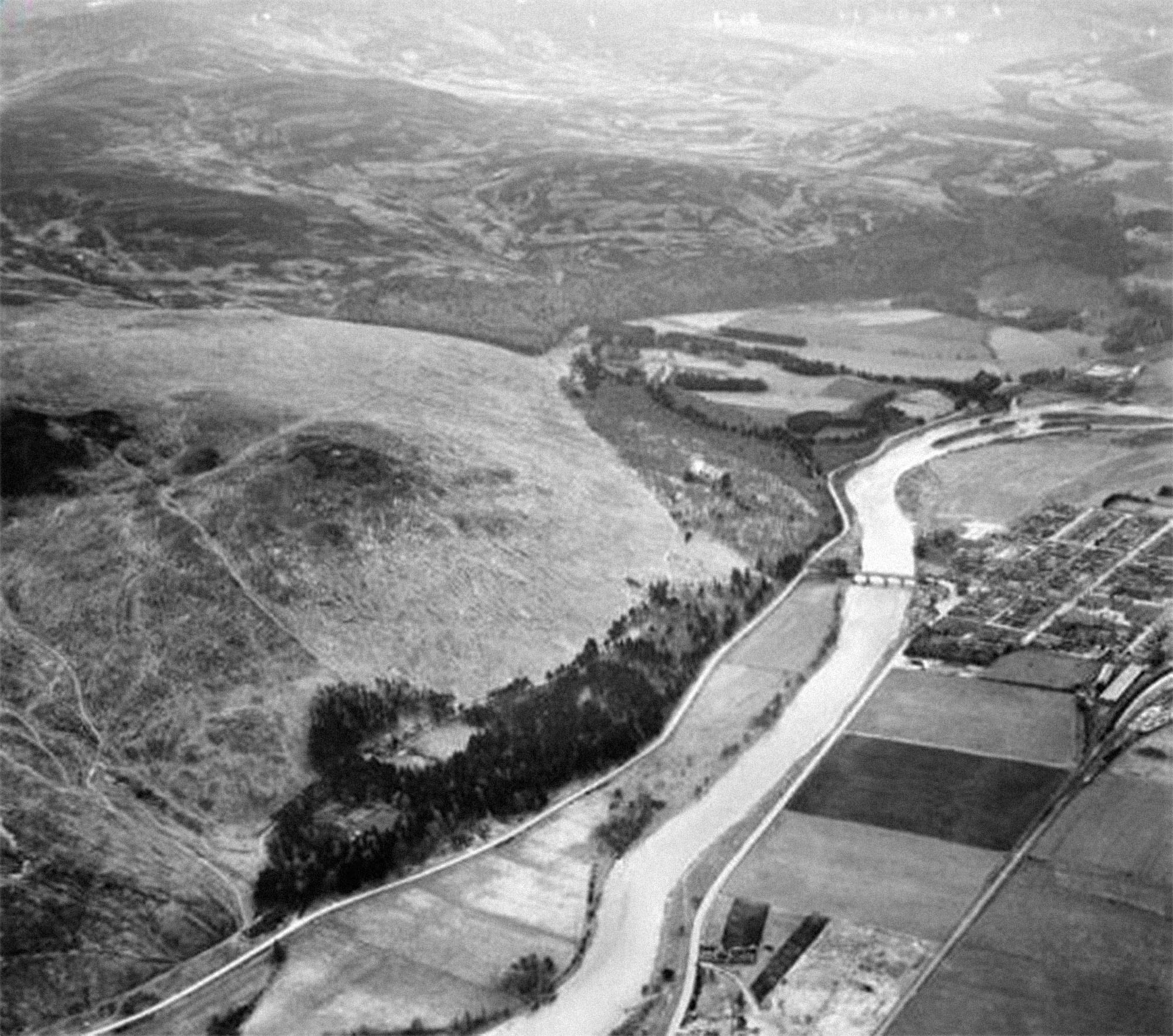 A black and white aerial photograph of denuded rolling hills on the left side of a wide river and fields and streets on the right.