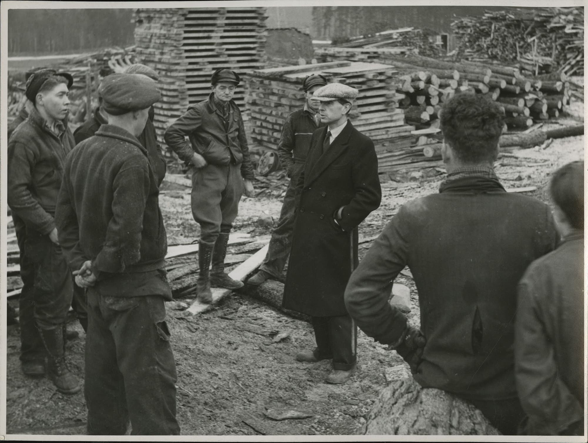A black and white photograph of a handful of men in work clothes standing in a half-circle facing a man in a dark overcoat and grey cap. The commissioner’s hands are in his coat pockets, a cane hangs from his left wrist. Logs and wooden pallets are stacked in the background.